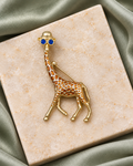Blue Shade Safari Brooch: gold-tone giraffe with blue sunglasses, a hat and amber-colored spots on ivory stone and sage satin