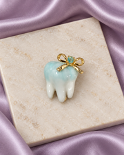 Dental Gleam Brooch with a softly aqua-tinted tooth motif and gold-tone bow, styled on ivory stone with lavender satin