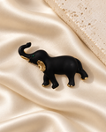 Midnight Tusker Brooch, a black elephant with gold-tone details and a green eye, styled flat on ivory satin
