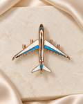 Skyline Jet Brooch: warm gold-tone airplane with white and turquoise-blue wings on ivory stone and champagne satin
