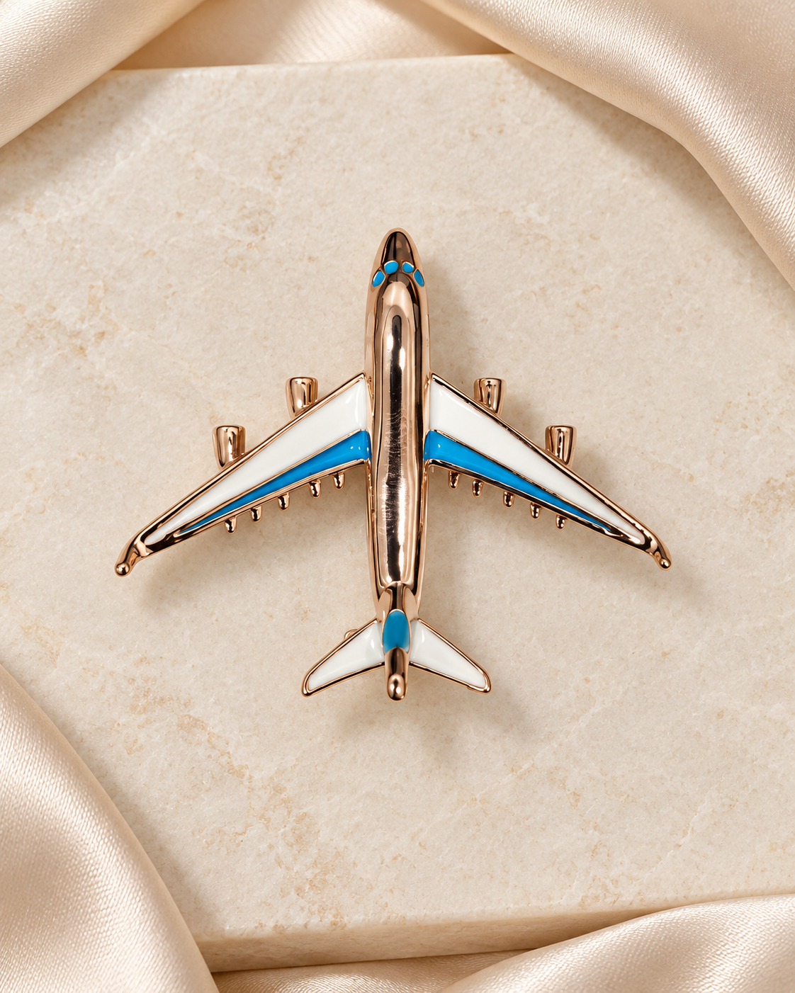 Skyline Jet Brooch: warm gold-tone airplane with white and turquoise-blue wings on ivory stone and champagne satin