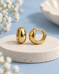 The Bare Glow Hoops with a smooth polished gold-tone finish on an ivory pedestal with a powder-blue background