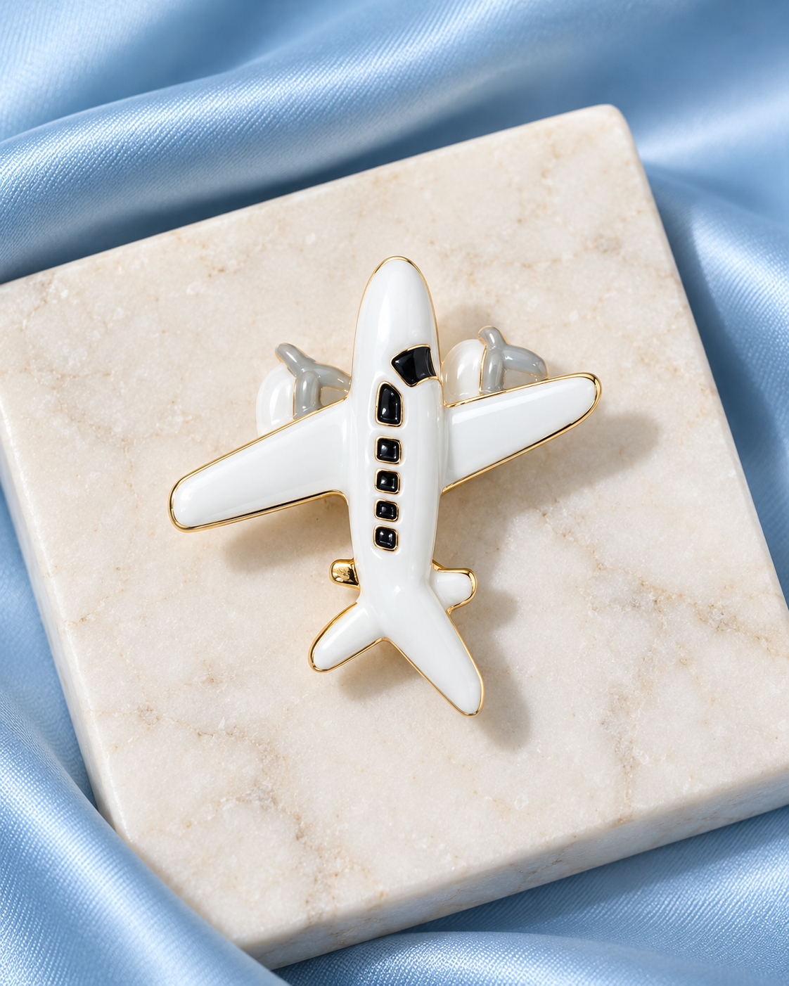 Wanderwing Brooch: glossy white airplane with black windows and gold-tone edging on sky-blue satin and ivory stone